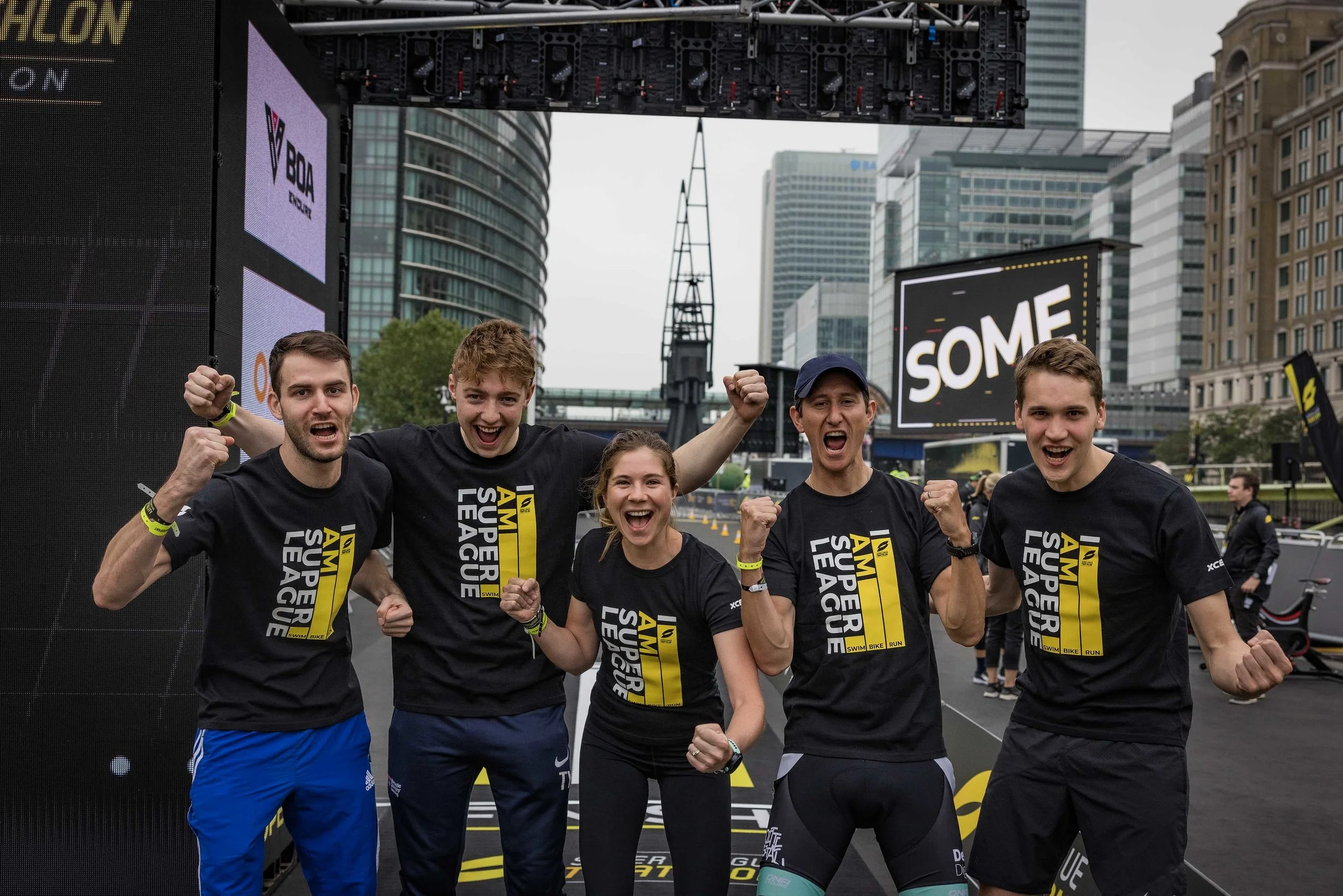 Group of five young people celebrating after a race, wearing black t-shirts with 'AM SUPPER LEAGUE' printed on them, in an urban outdoor setting with tall buildings and a large digital screen background.