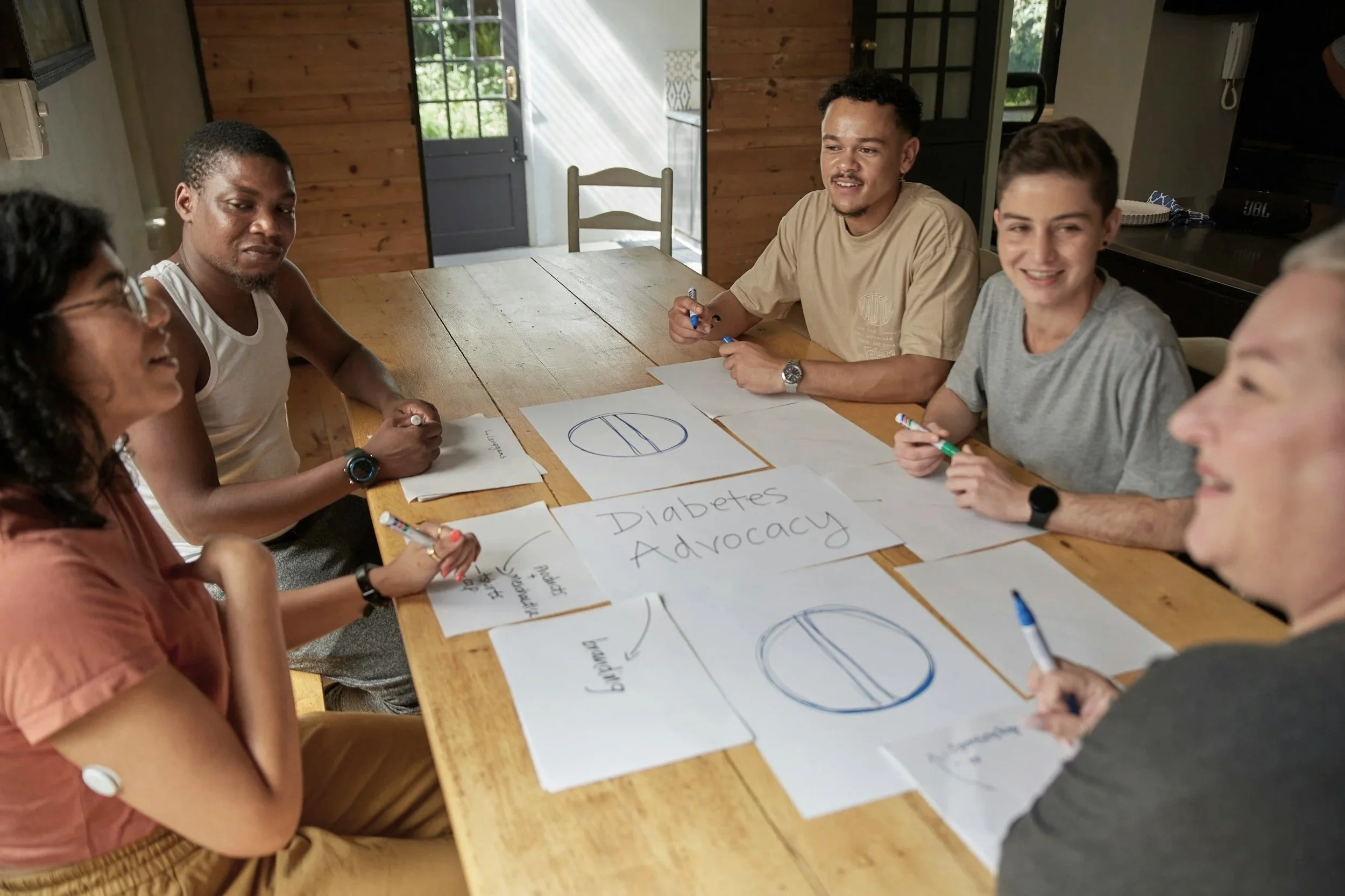Group of six diverse people sitting around a wooden table in a room, engaged in a meeting or discussion about diabetes advocacy, with papers and diagrams on the table.