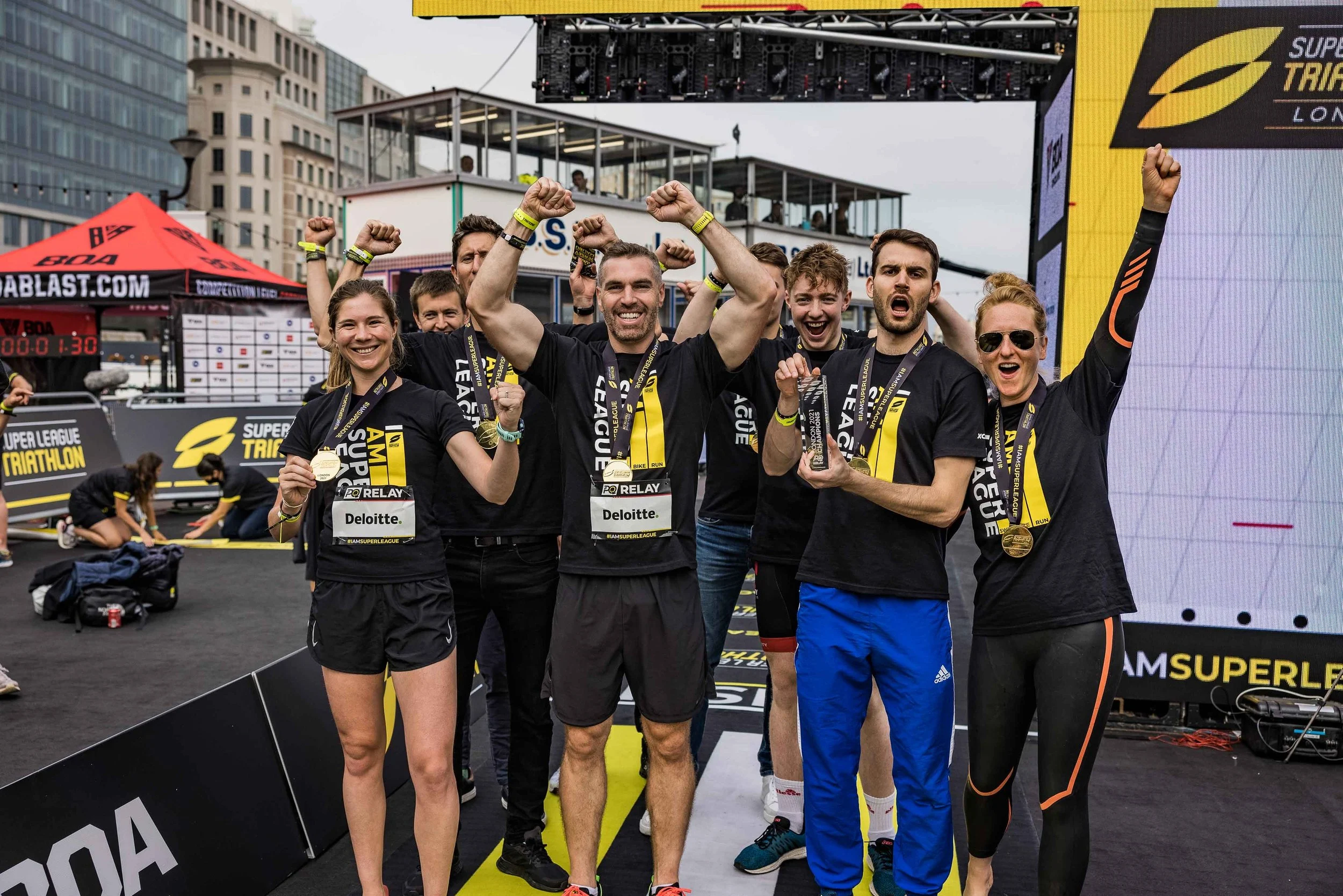 A group of athletes celebrating with medals and a trophy at a triathlon event, standing on a stage with a large digital screen and city buildings in the background.