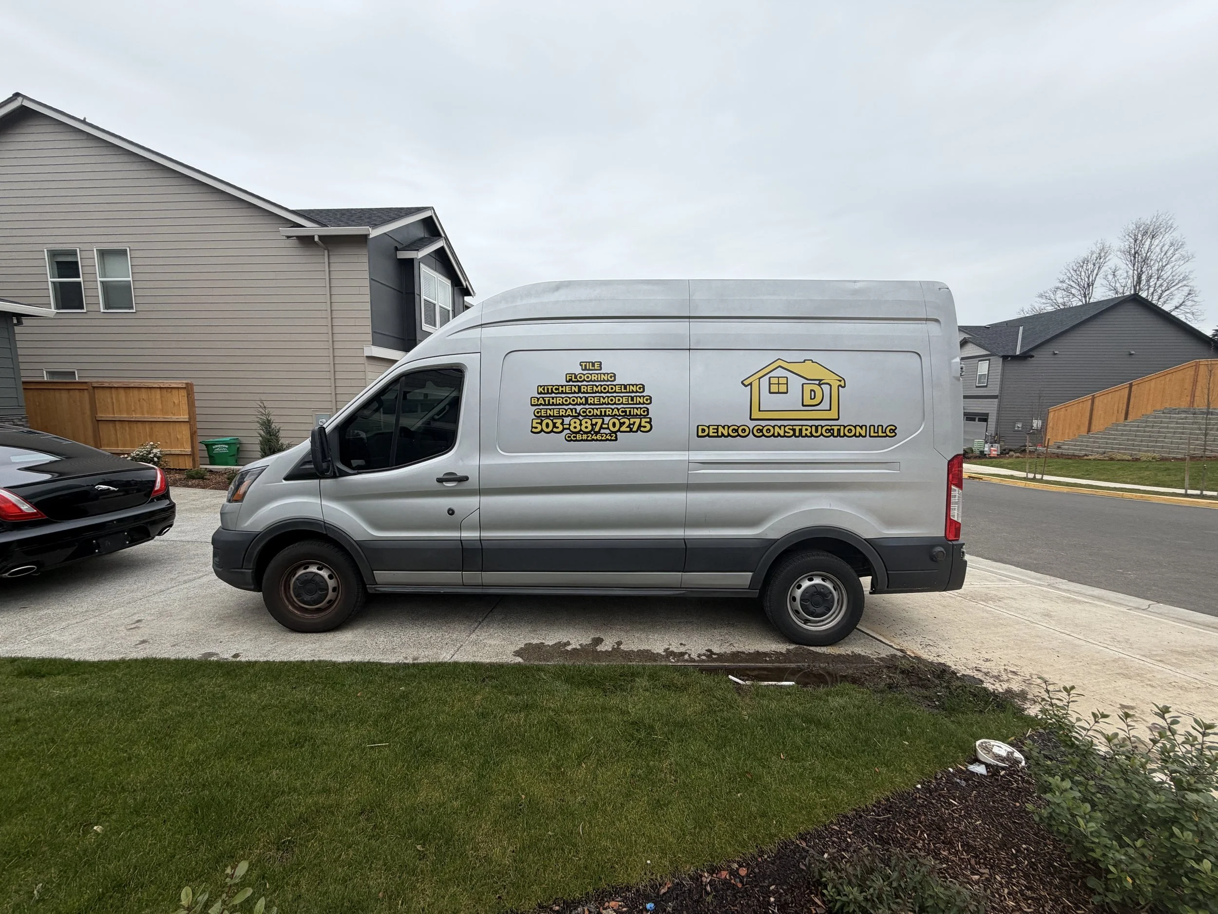 A white work van parked in front of residential houses, with services listed on the side, including tile, flooring, kitchen remodeling, bathroom remodeling, general contracting, along with a phone number and company name Denco Construction LLC.
