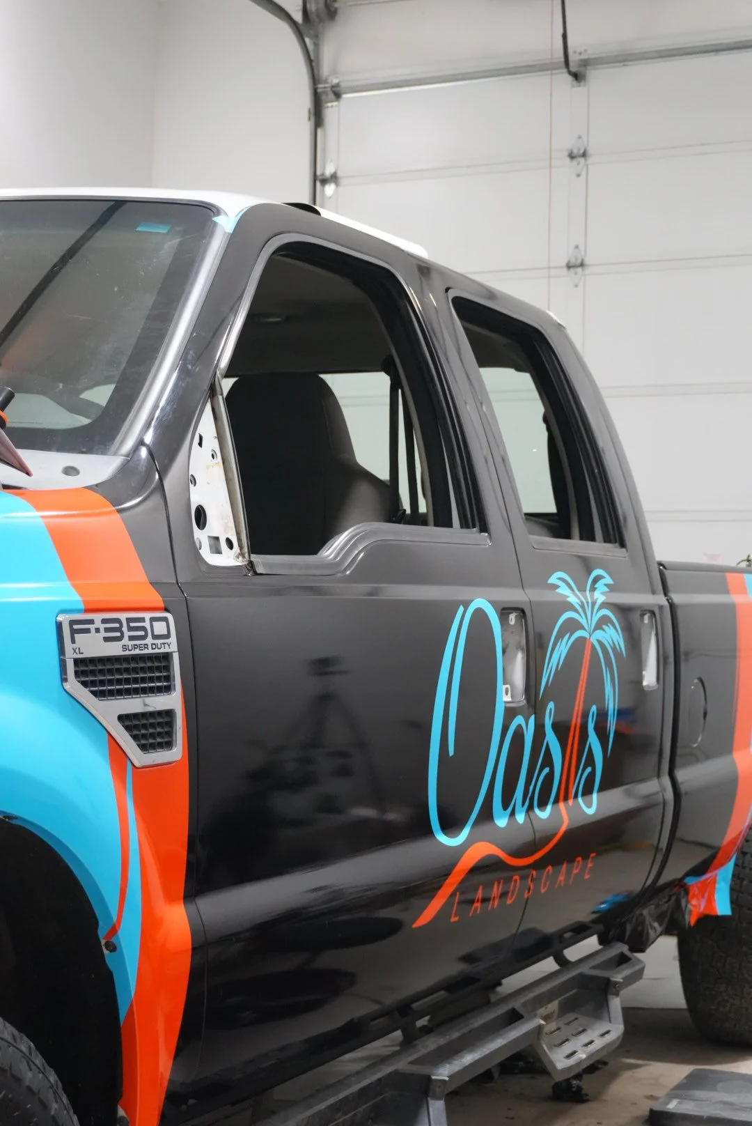 A black pickup truck with a large logo on the side that says 'Oasis Landscape' in stylized blue and orange text, inside a garage.