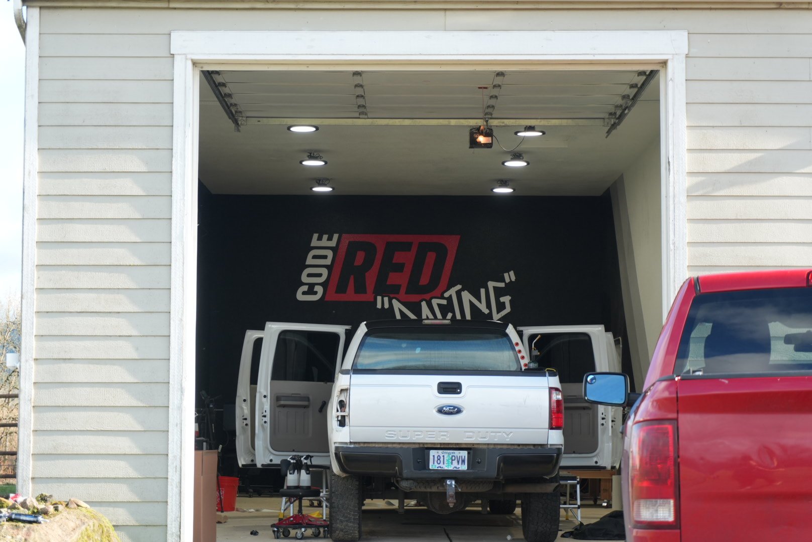 A garage with an open door, displaying a white Ford Super Duty pickup truck with its tailgate down, and orange lights on the ceiling inside. A red vehicle is partially visible at the right edge of the image. The garage has a black wall 