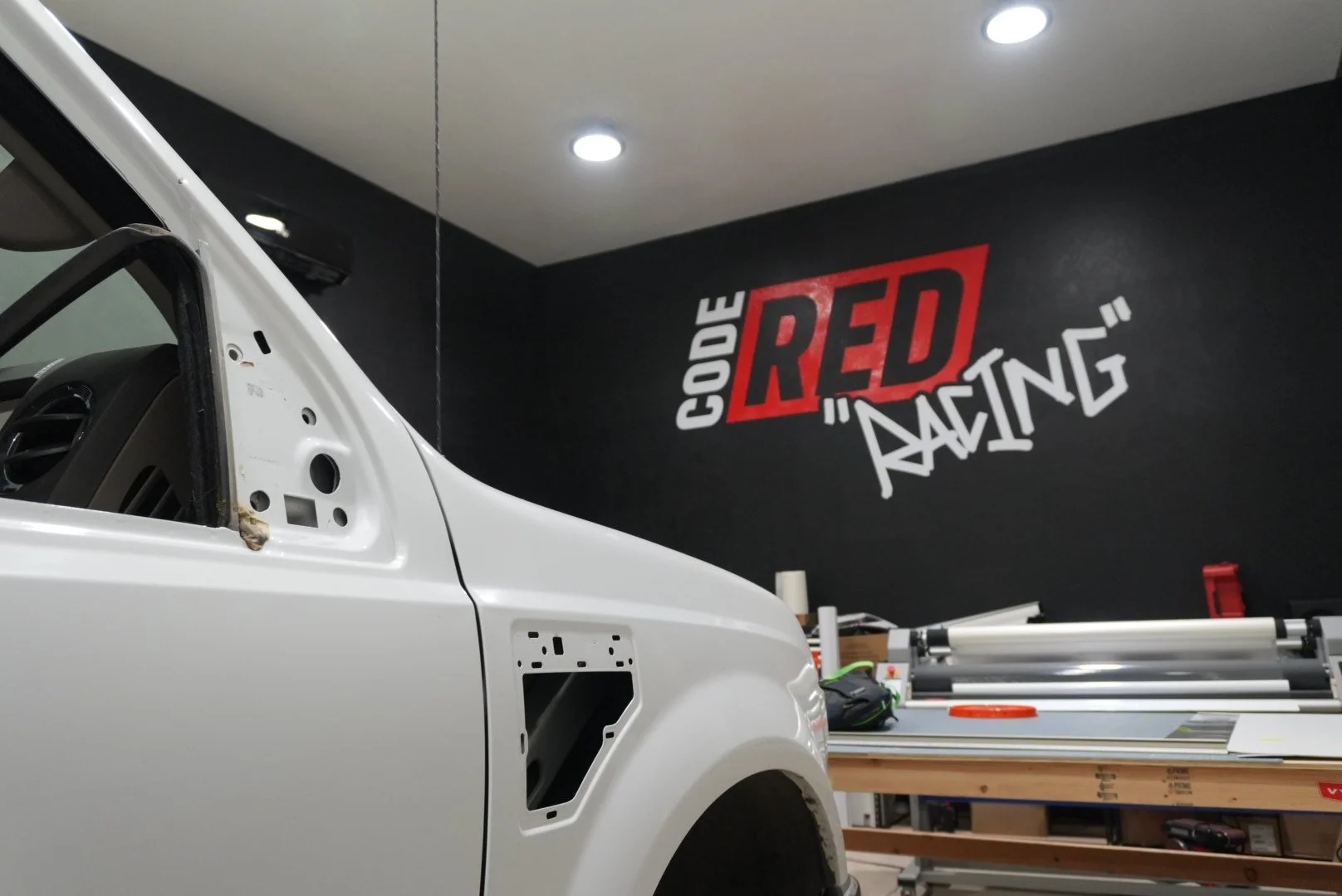 Partially assembled white race car in a workshop with a black wall featuring the 'Code Red Racing' logo in red and white.