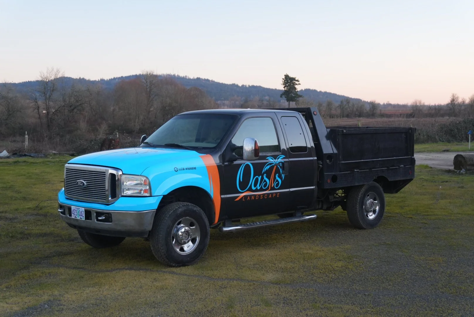 A pickup truck with a black bed and a colorful wrap that says 'Oasis Landscape' parked on grass with trees and hills in the background.