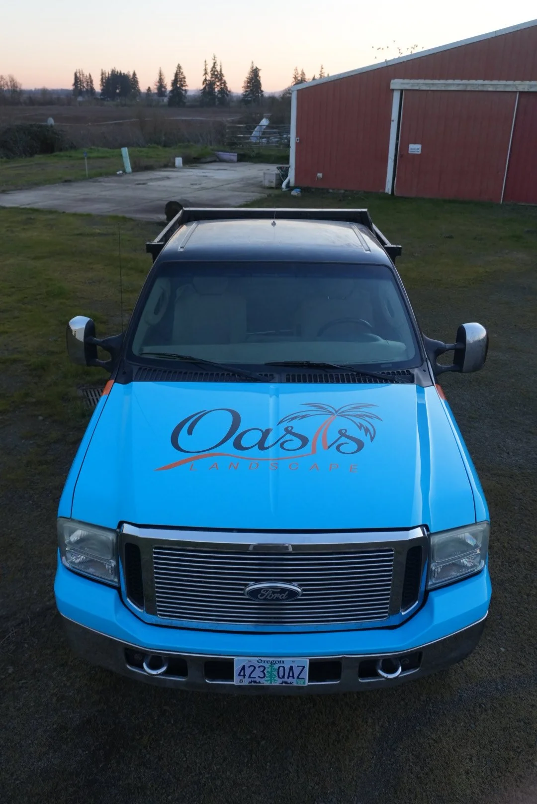 Front view of a blue Ford truck with 'Oasis Landscape' logo and palm tree graphic on the hood, parked on a grassy yard near a red barn and open farmland.