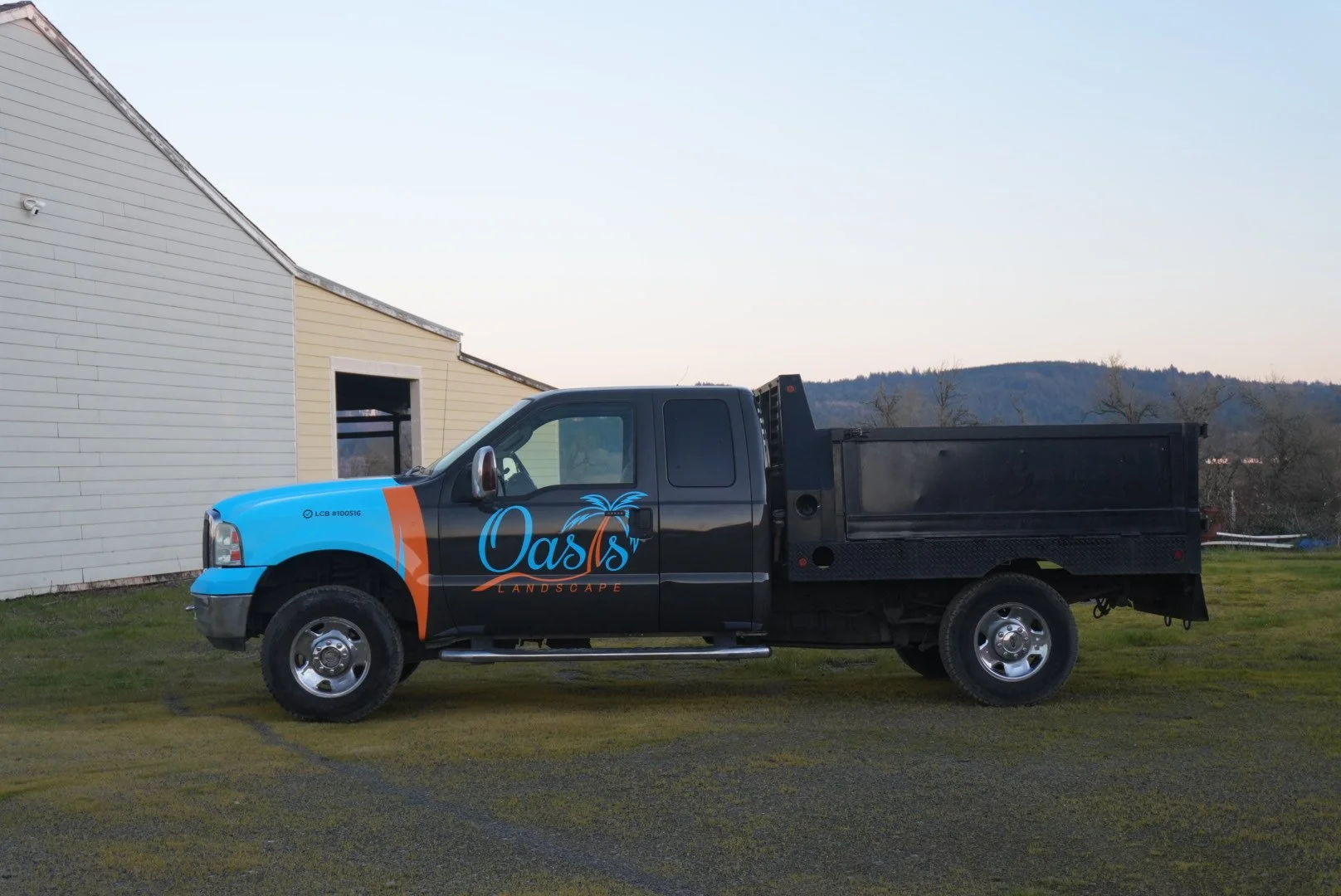 A black and blue pickup truck with a city logo that reads 'Oasis Landscape' parked outside near a building with a gray and beige exterior and a grassy area, with hills in the background.