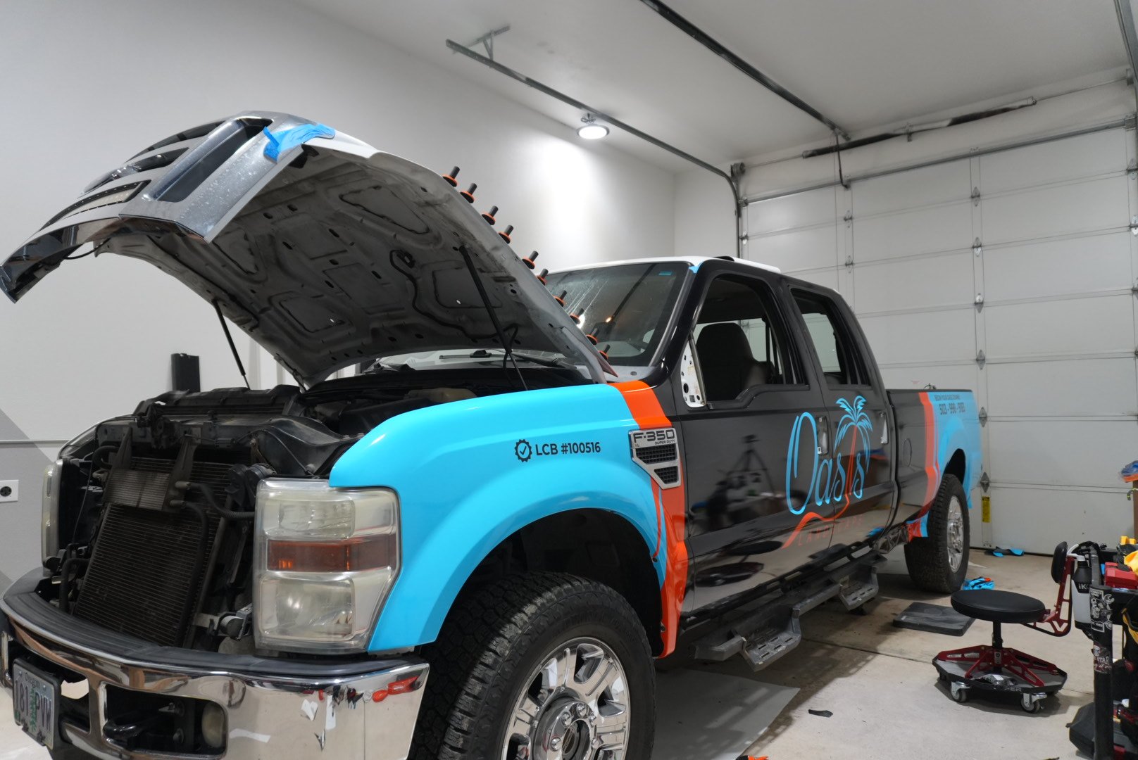 A pickup truck in a garage with open hood, painted with a black, blue, and orange design and the word 'Oasis' on the side, surrounded by tools and equipment.