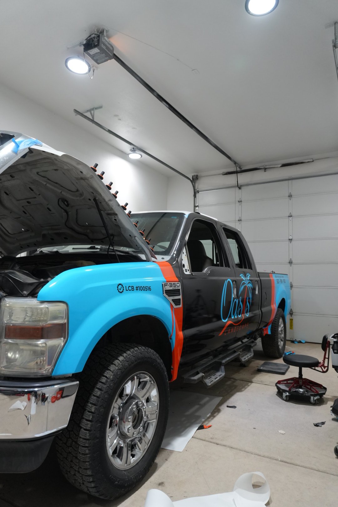 A truck inside a garage with its hood open, decorated with a custom wrap that features a tropical island theme, including a palm tree and the word 'Oasis'.