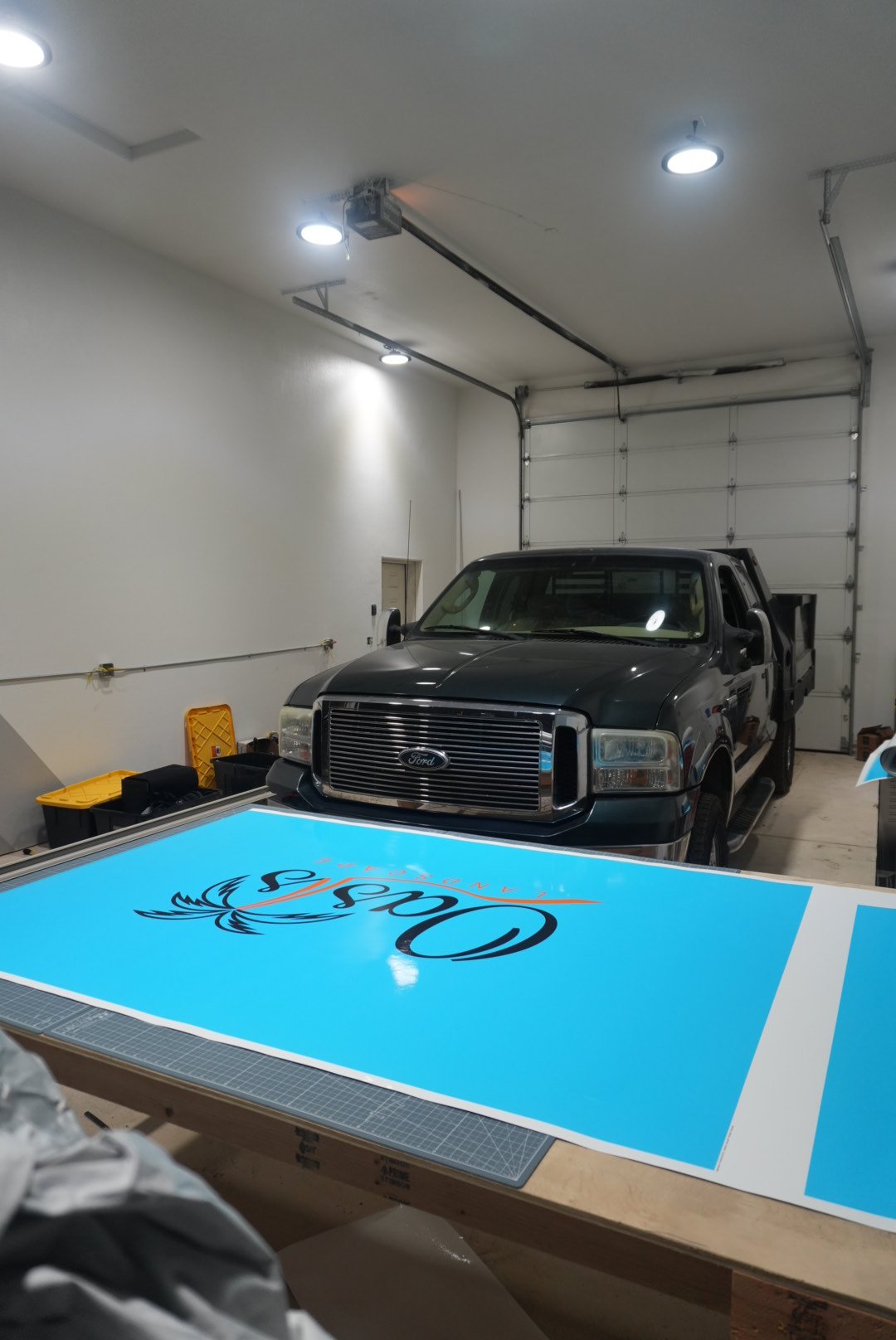 Garage with a black Ford truck parked in front of a closed garage door and a large sign with the text 'OASIS' on a work table in the foreground.