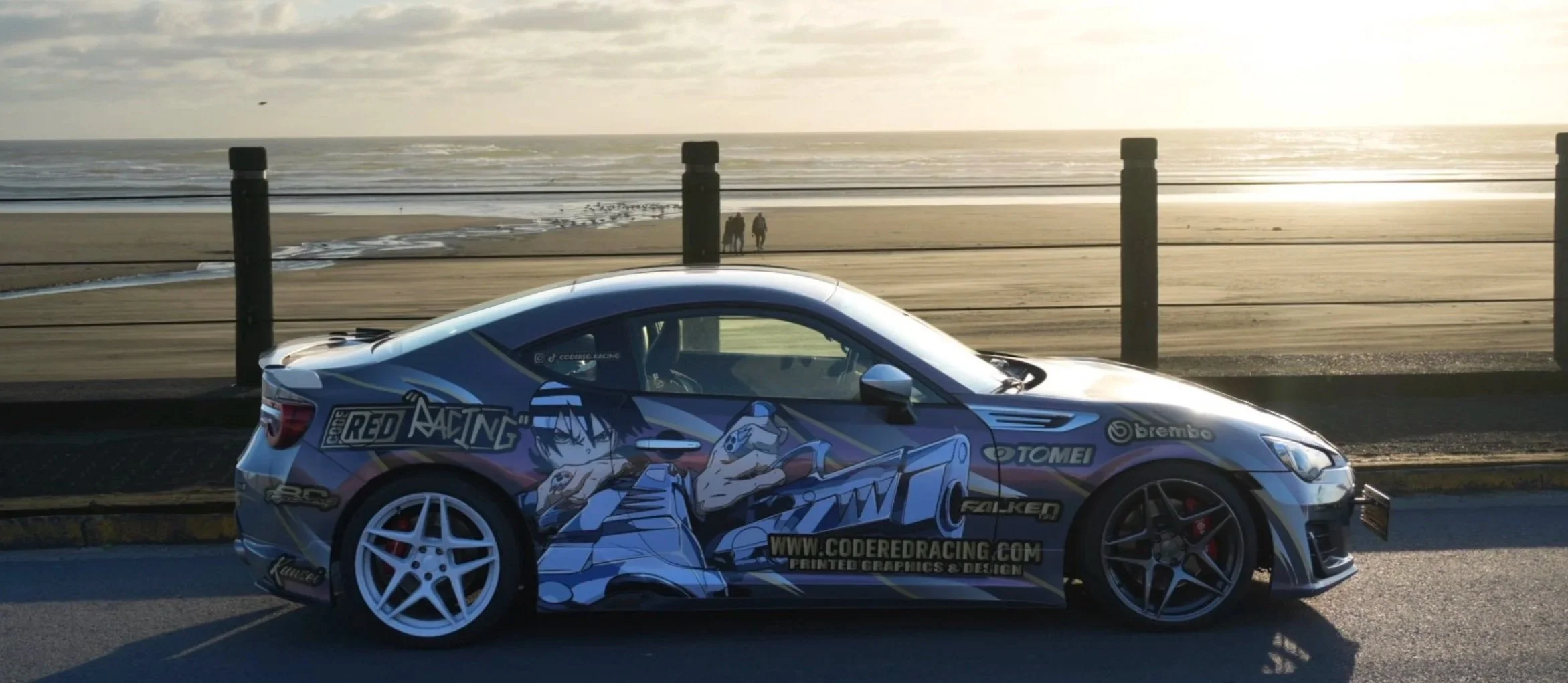 A race car with anime-style artwork wrap and sponsorship logos sticker parked along a beach boardwalk at sunset.