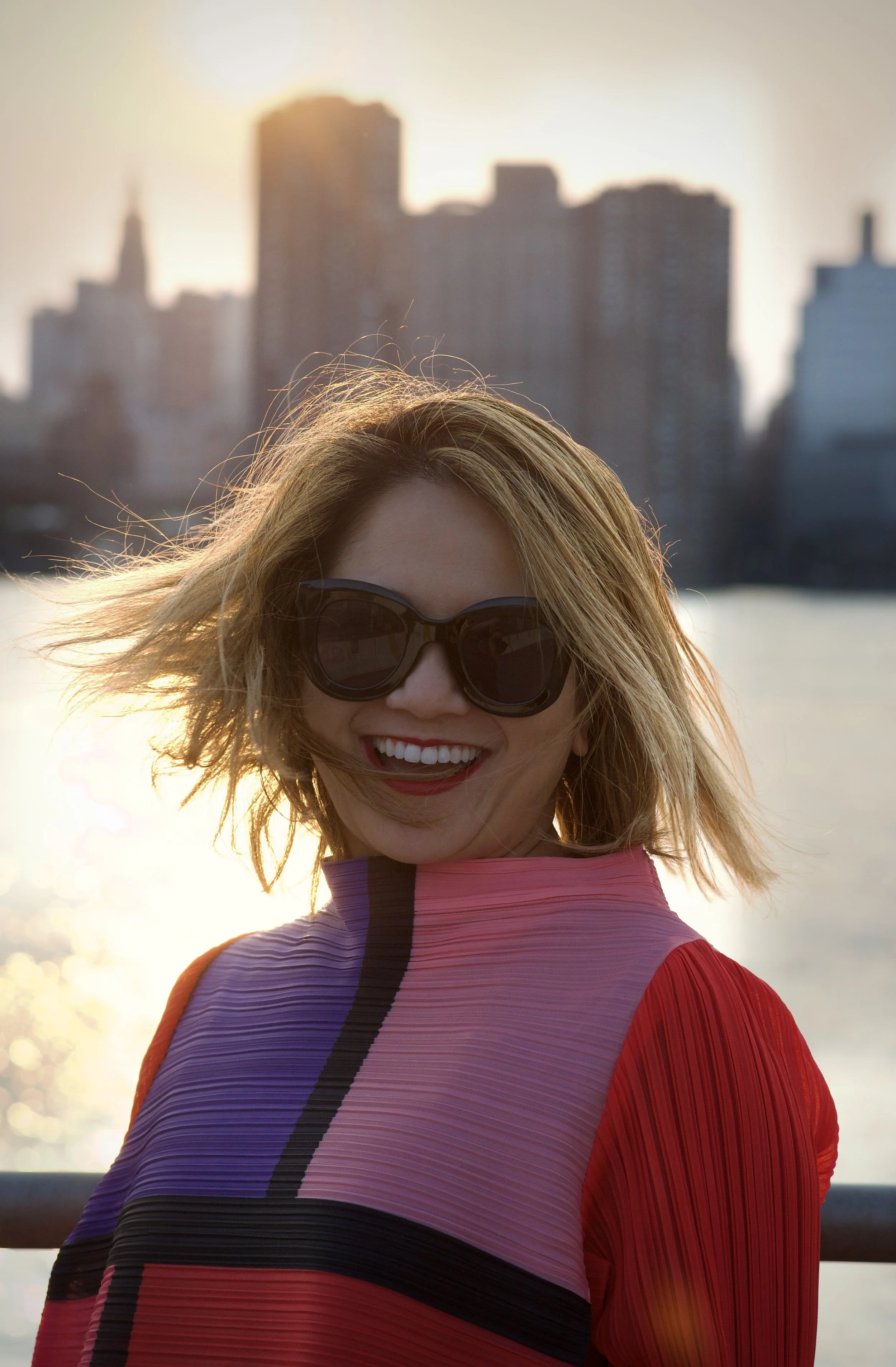A woman with blond hair wearing dark sunglasses, smiling, dressed in a colorful striped top, standing outdoors with a city skyline and water in the background during sunset.