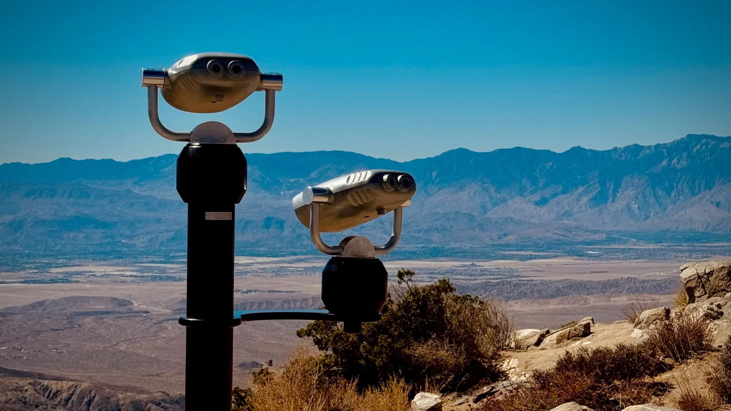Two coin-operated binocular viewers overlooking a desert landscape with mountains in the background.