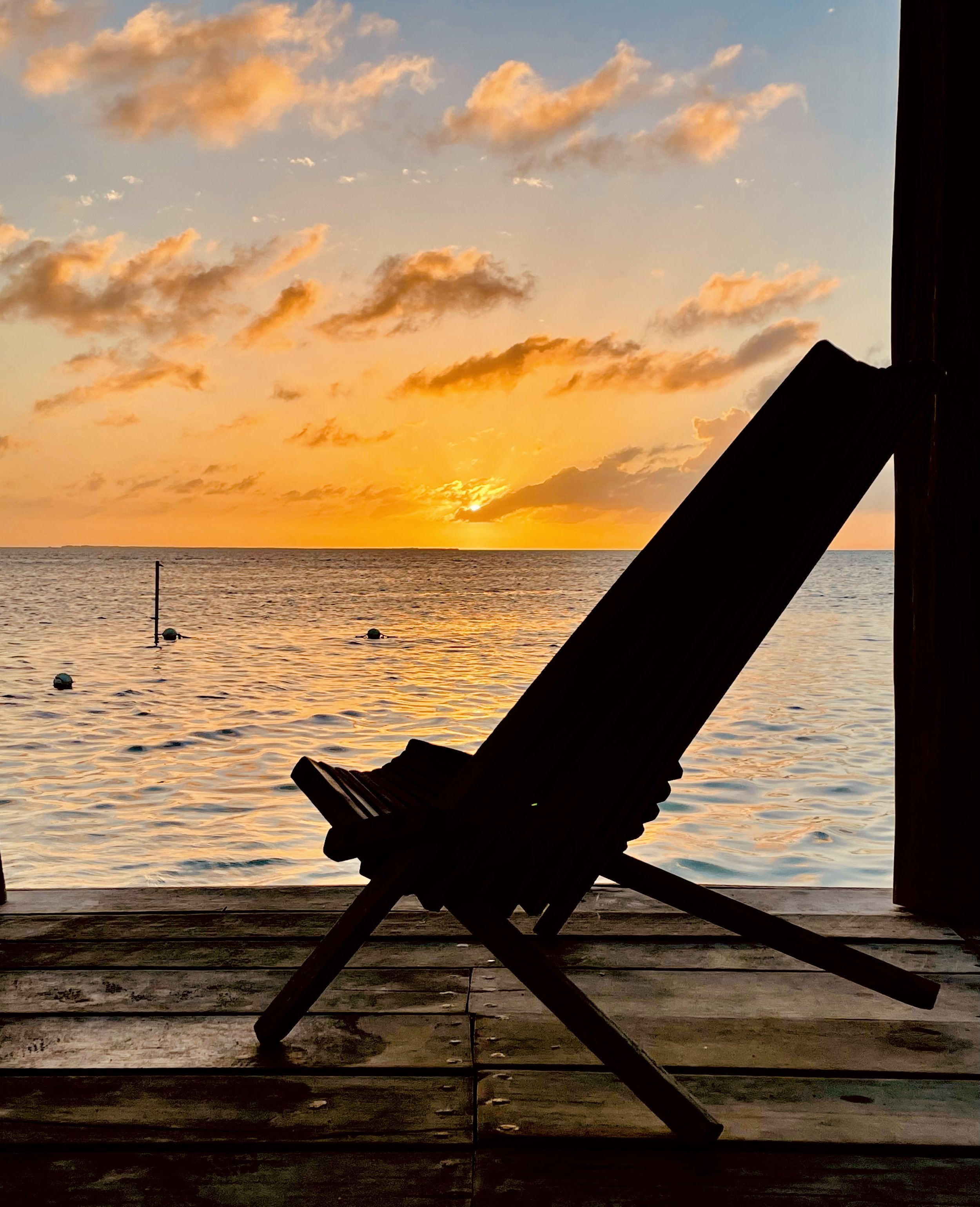 Silhouette of a folding chair on a wooden deck facing a sunset over the ocean, with clouds in the sky, captured from inside a structure with dark framing on the right.