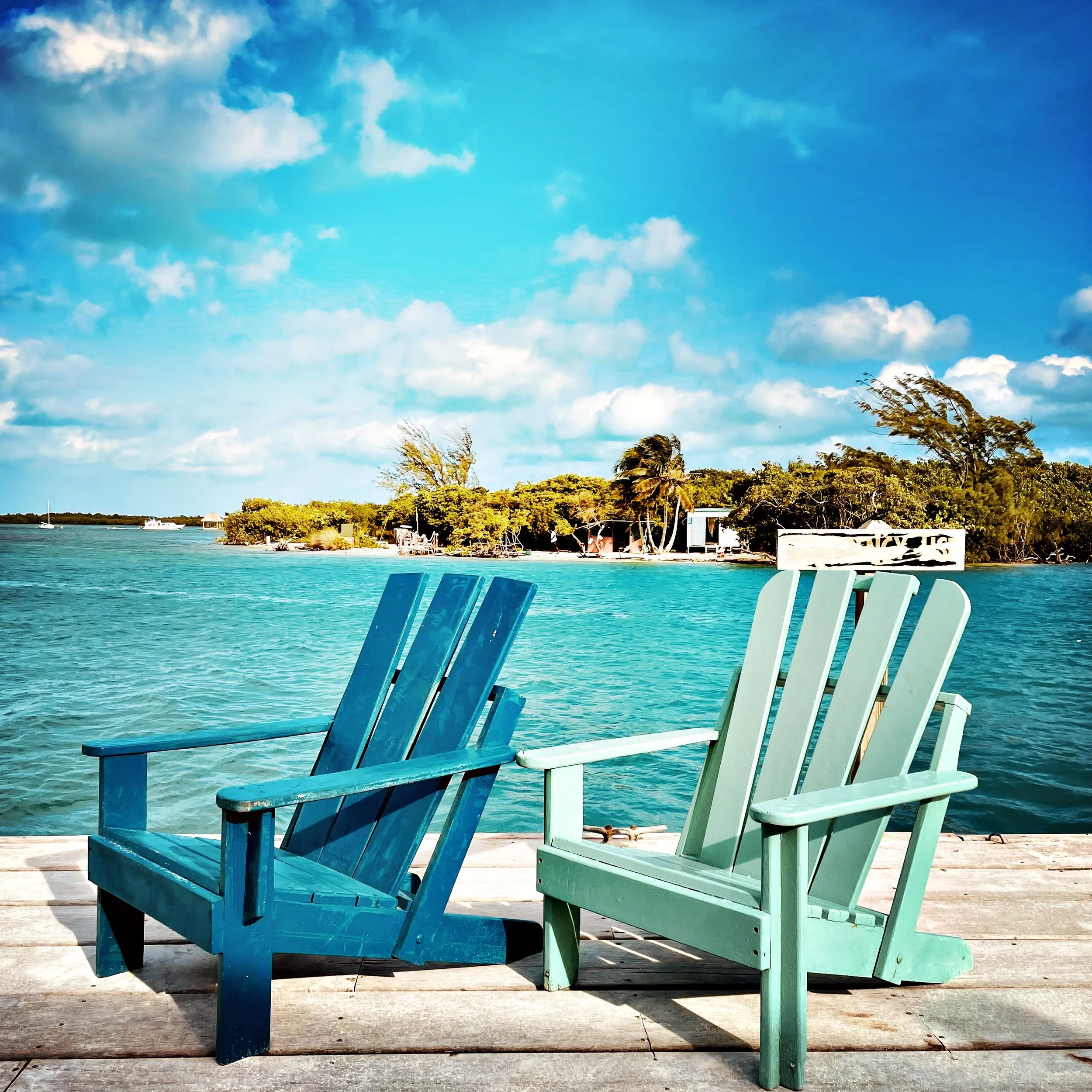 Two Adirondack chairs, one painted blue and the other painted mint green, are positioned on a wooden dock near a body of turquoise water. In the background, there are trees, some of which are swaying in the wind, and a partly cloudy sky with bright s