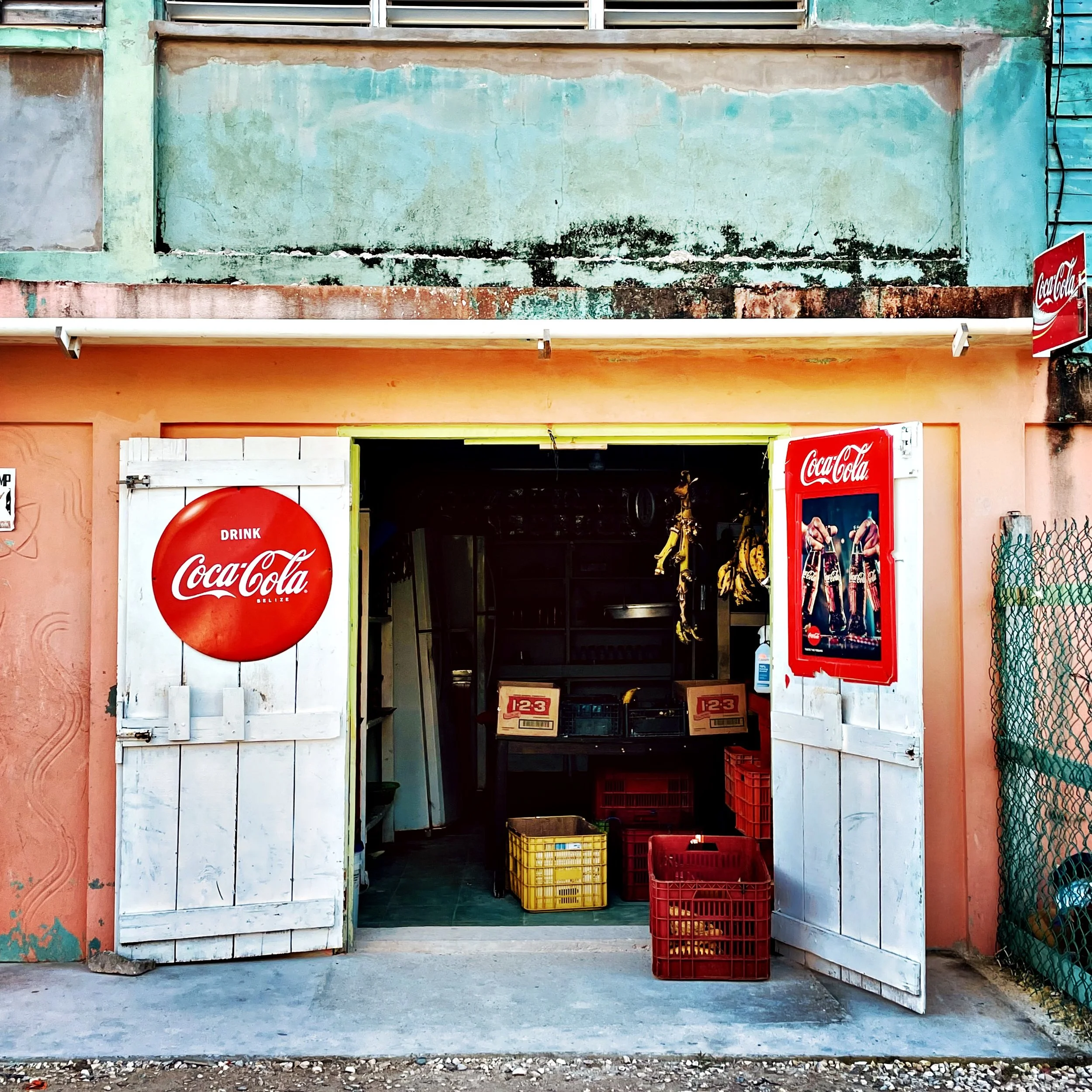 Open storefront with white wooden door, Coca-Cola sign, yellow and red plastic crates, and shelves inside. Pink wall and chain-link fence outside.