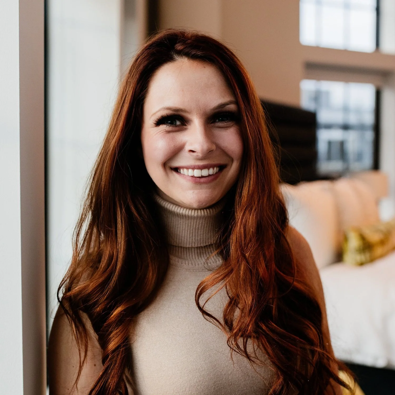 Woman with long red hair smiling indoors near a window.