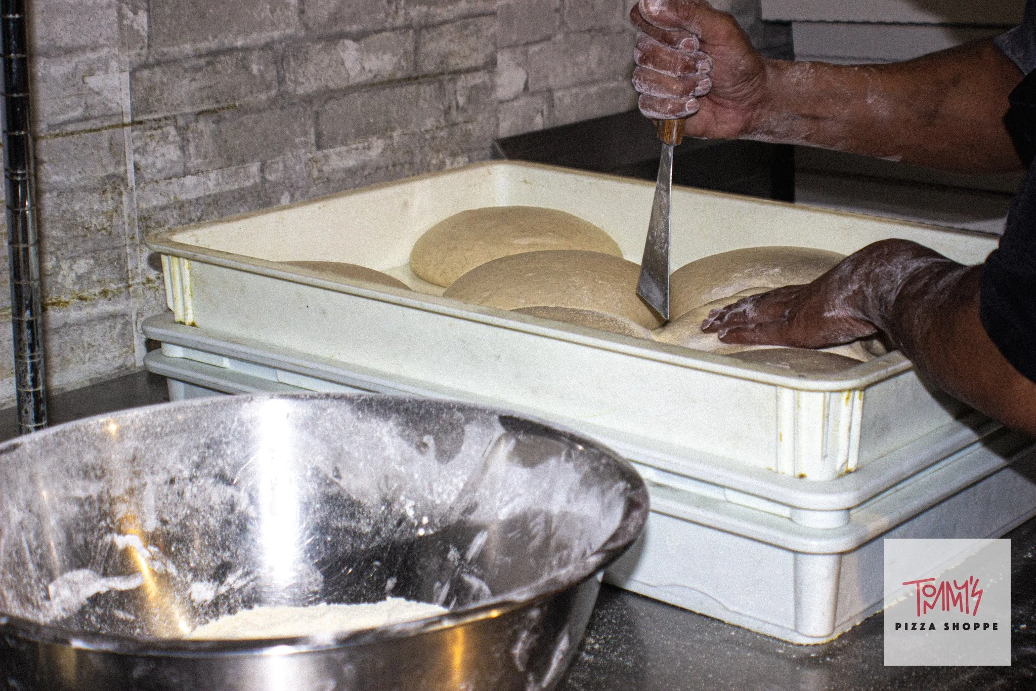 A person mixing dough in a large metal bowl in a kitchen, with a dough cutter and a plastic container with dough in the background.