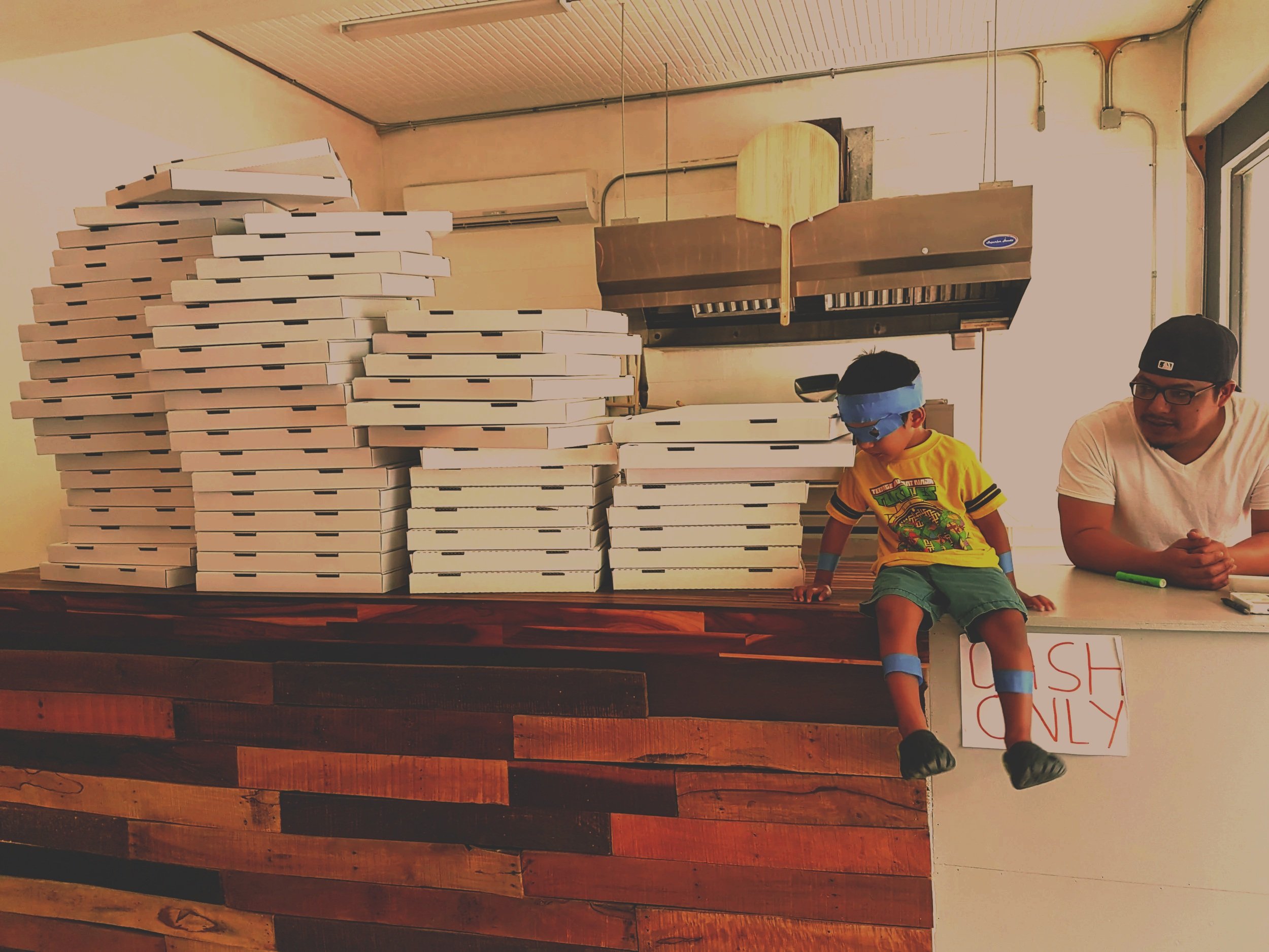 A man and a young boy sitting at a table with a large stack of pizza boxes in front of them indoors. The man is wearing glasses and a cap, and the boy is wearing a dinosaur t-shirt, shorts, and a blue blindfold. The background includes an air conditioning unit, visible electrical wiring, and a wall with a window.