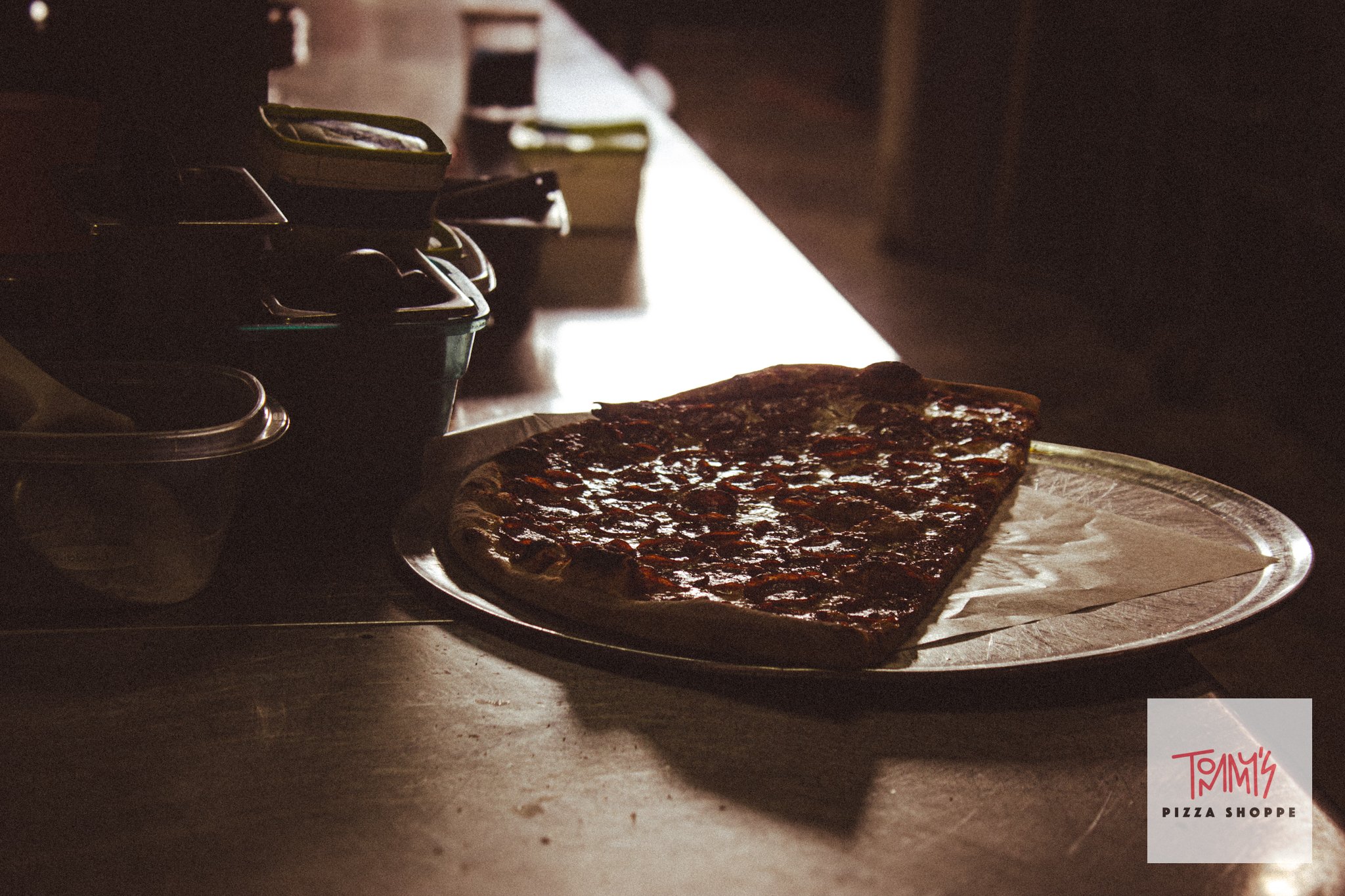 A pizza with pepperoni slices on a paper-lined tray in a dimly lit room, with a box of Tommy's Pizza Shoppe visible in the bottom right corner.