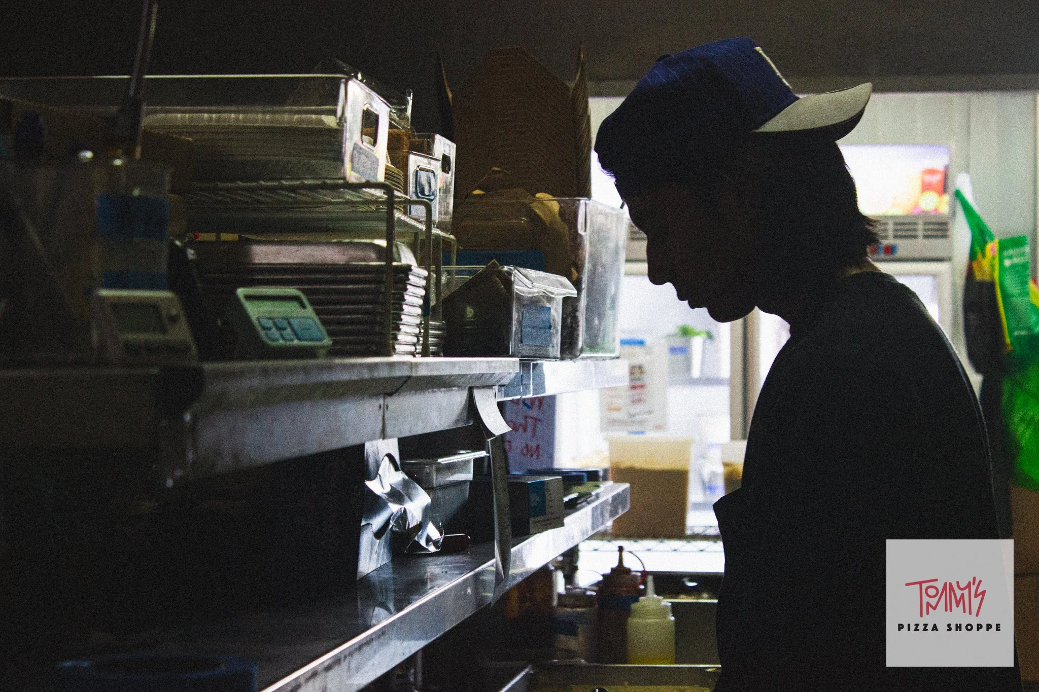 A silhouette of a person working behind the counter at a pizza shop, with shelves of supplies and a menu board in the background.