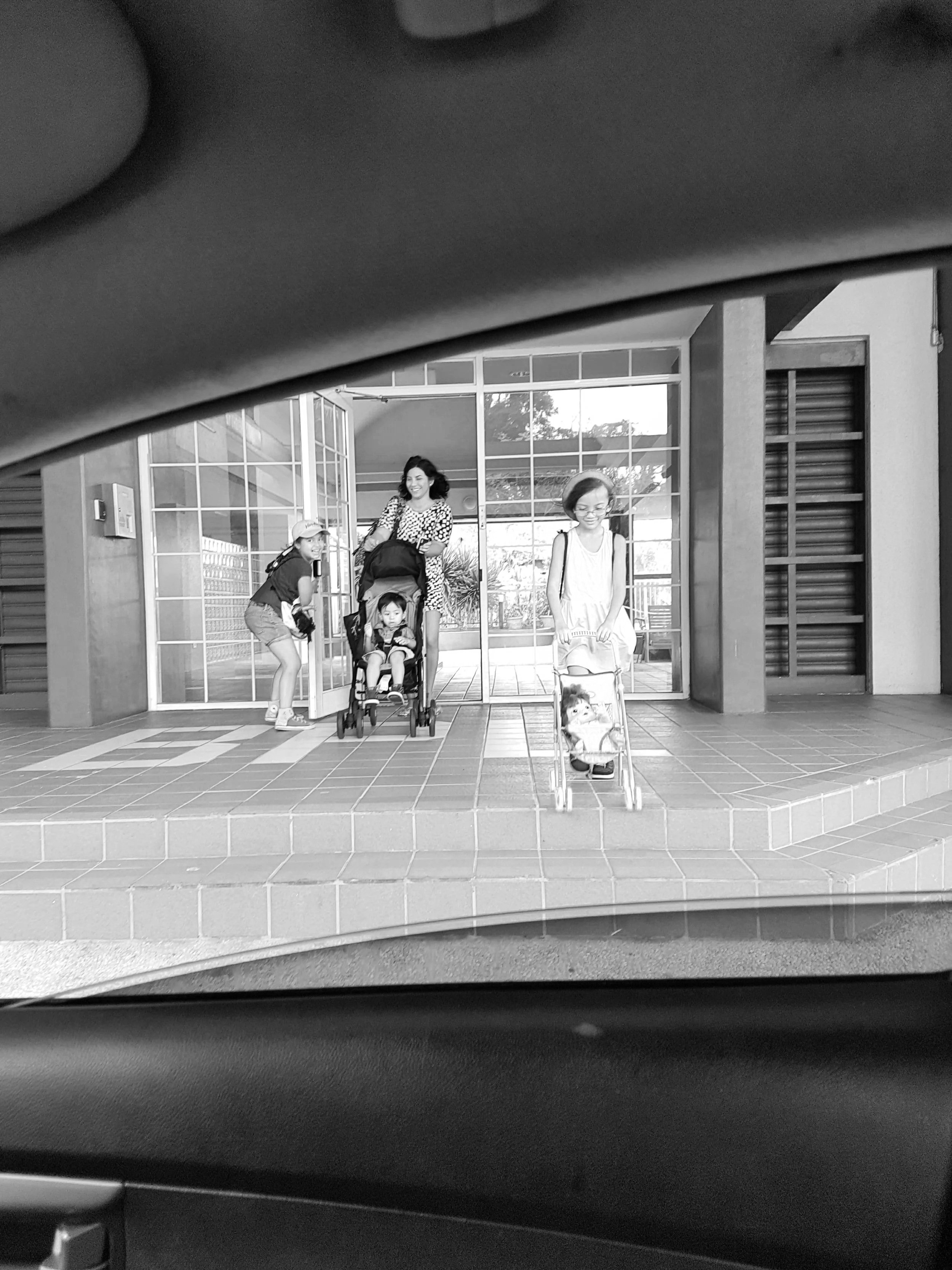 People exiting a building through glass doors, with a woman and children, one in a stroller, and one riding a scooter, seen from inside a vehicle.