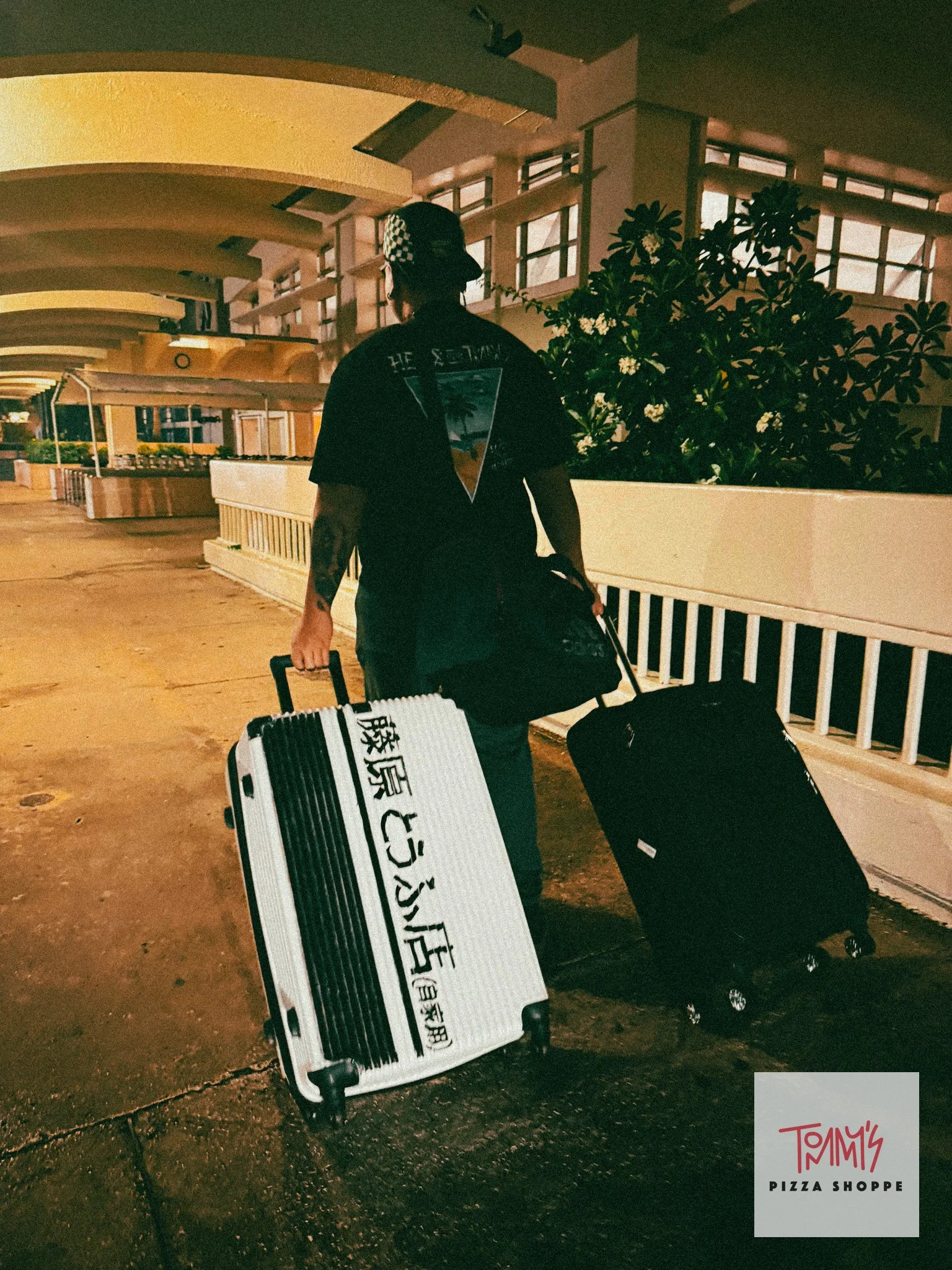 A person walking through an outdoor area at night, pushing two rolling suitcases. The man is wearing a dark shirt and a cap with a checkered pattern. There are buildings with large windows in the background and some plants. A sign for Tommy's Pizza Shoppe is in the bottom right corner.