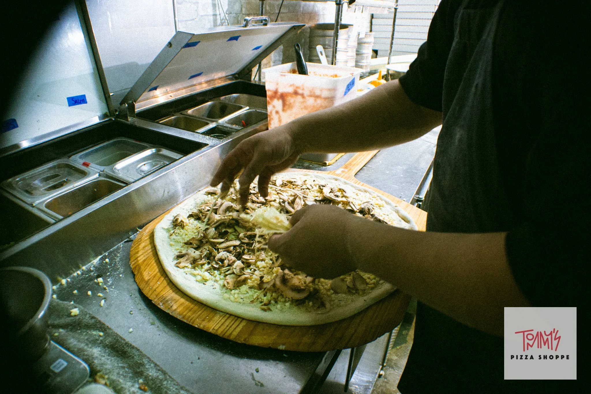 Person preparing a pizza with toppings such as mushrooms and cheese on a wooden pizza peel at a pizza shop, with a pizza shop logo labeled 'Tommy's Pizza Shoppe' visible in the bottom right corner.