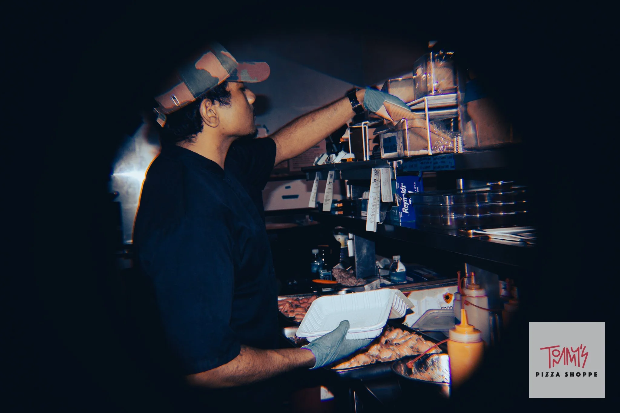 A person wearing a camouflage cap and gloves preparing food at a pizza shop counter. They are reaching into a food container, with pizza slices, bottles of condiments, and pizza shop branding visible on the counter.