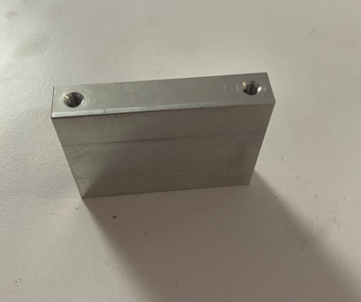 A metal rectangular block with two threaded holes on the top surface, placed on a light-colored surface.