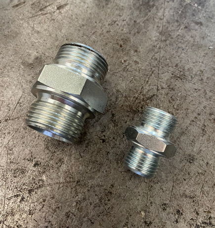 Two metal threaded fittings on a textured gray surface. The larger fitting is on the left and the smaller on the right.