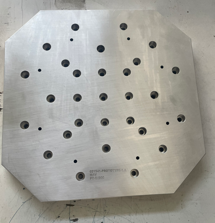 A large, flat, metallic square plate with multiple drilled holes of varying sizes.
