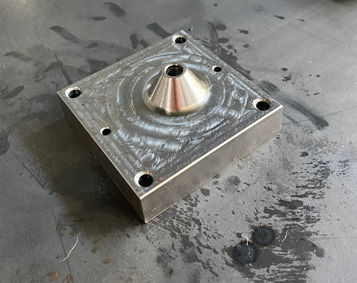 Metal workpiece with a conical feature and multiple drilled holes, placed on a work surface.