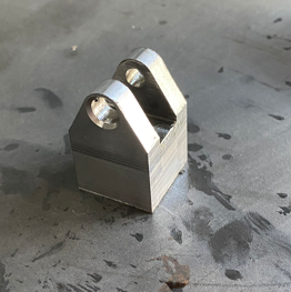 A small metallic component with two circular holes, placed on a workshop surface.