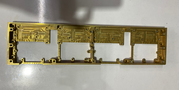 A long, rectangular, gold-colored electronic circuit board with multiple compartments and pathways.