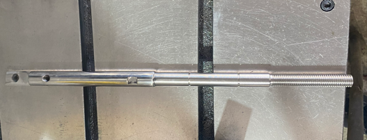 A metal rod with a hole and notches, lying on a metal surface with black support frames.