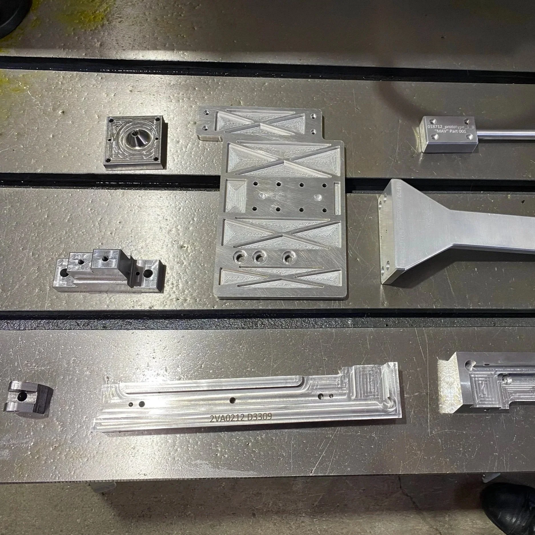 Several metal parts with various shapes and drilled holes are laid out on a workbench, including rectangular, L-shaped, and flat pieces, possibly for manufacturing or assembly purposes.