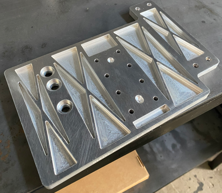 Metal workpiece with engraved geometric patterns and multiple drilled holes, placed on a black work surface.