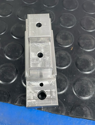 A close-up of a metal bracket with three drilled holes, sitting on a textured rubber mat.