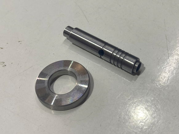 Metal machined components, one cylindrical part with grooves and a smaller ring-shaped piece, on a plain white surface.