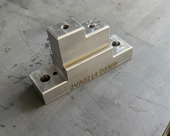 Metal mechanical component with three mounting holes and engraved identification codes, placed on a gray surface.