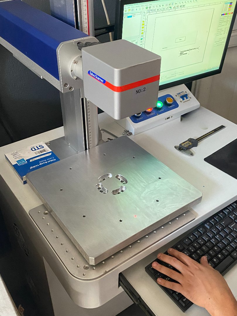 A coordinate measuring machine with a metal part on the measuring table, a computer monitor displaying a diagram or measurement software, and an empty keyboard. A person's hand is on the keyboard.