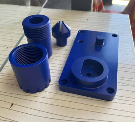 Blue plastic electrical or mechanical components on a wooden surface, including a cylindrical piece, a square piece with a cylindrical protrusion, and a smaller conical piece.