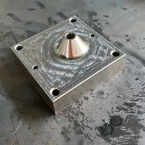 Metal machined block with a conical feature and surrounded by six drilled holes on a workbench.