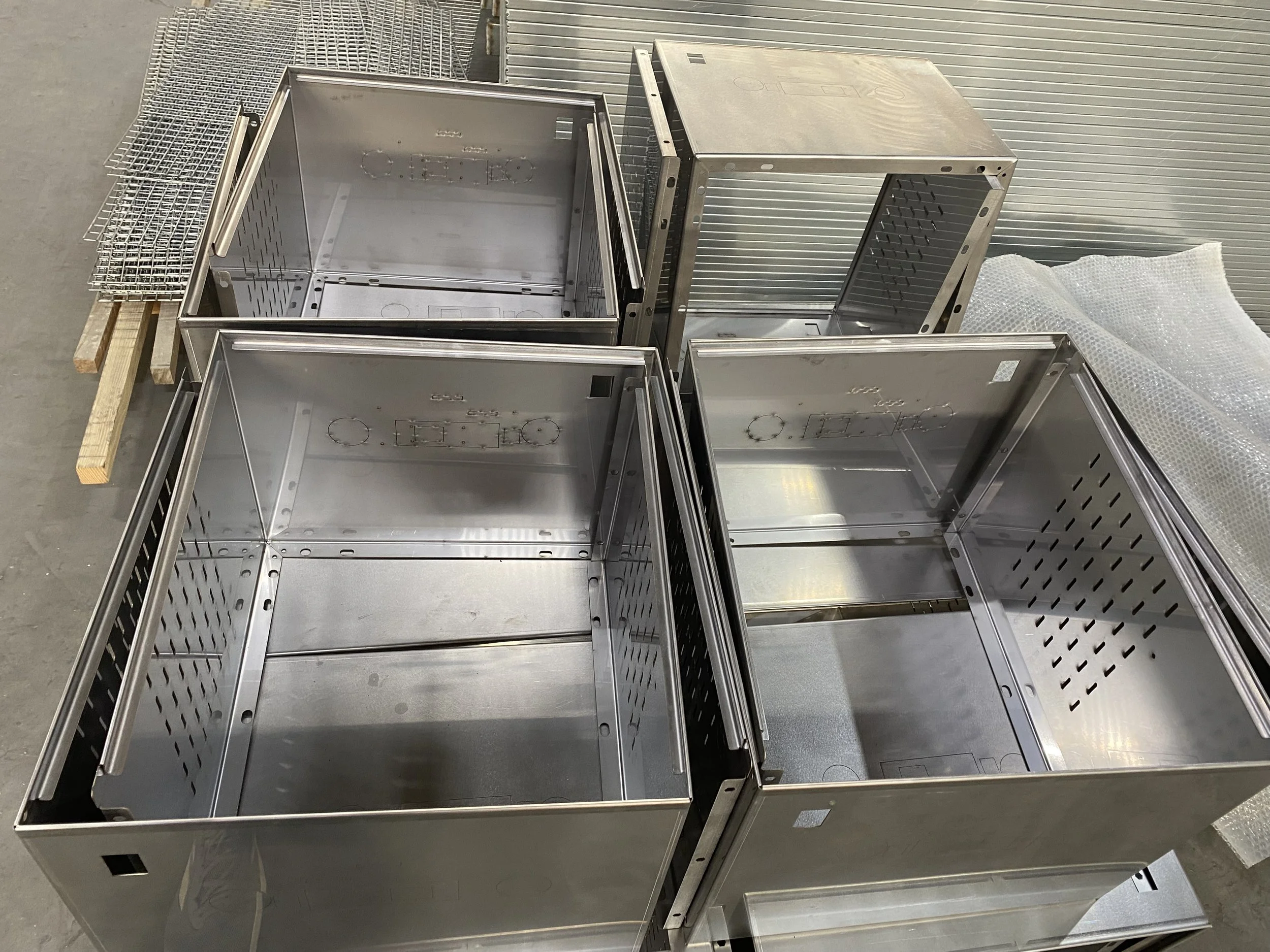 Four empty stainless steel enclosures or cabinets, some with ventilation holes, placed on a concrete floor in an industrial or manufacturing setting.