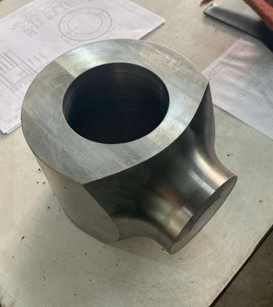 Metal machined part with a cylindrical hole on a work table, with technical drawings and papers in the background.