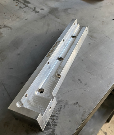 Metal machined part with holes and grooves, labeled 'GCC ECU 2007', placed on a workbench in a workshop.