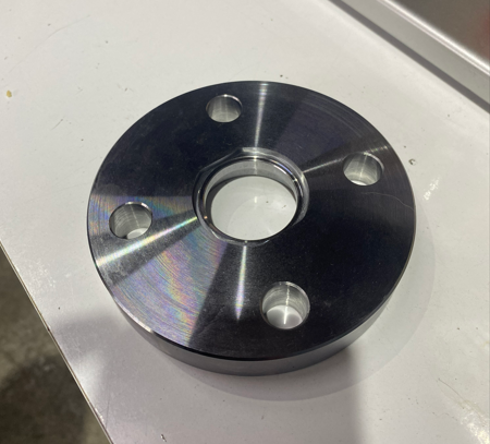 A round metal flange with four holes, placed on a white surface.