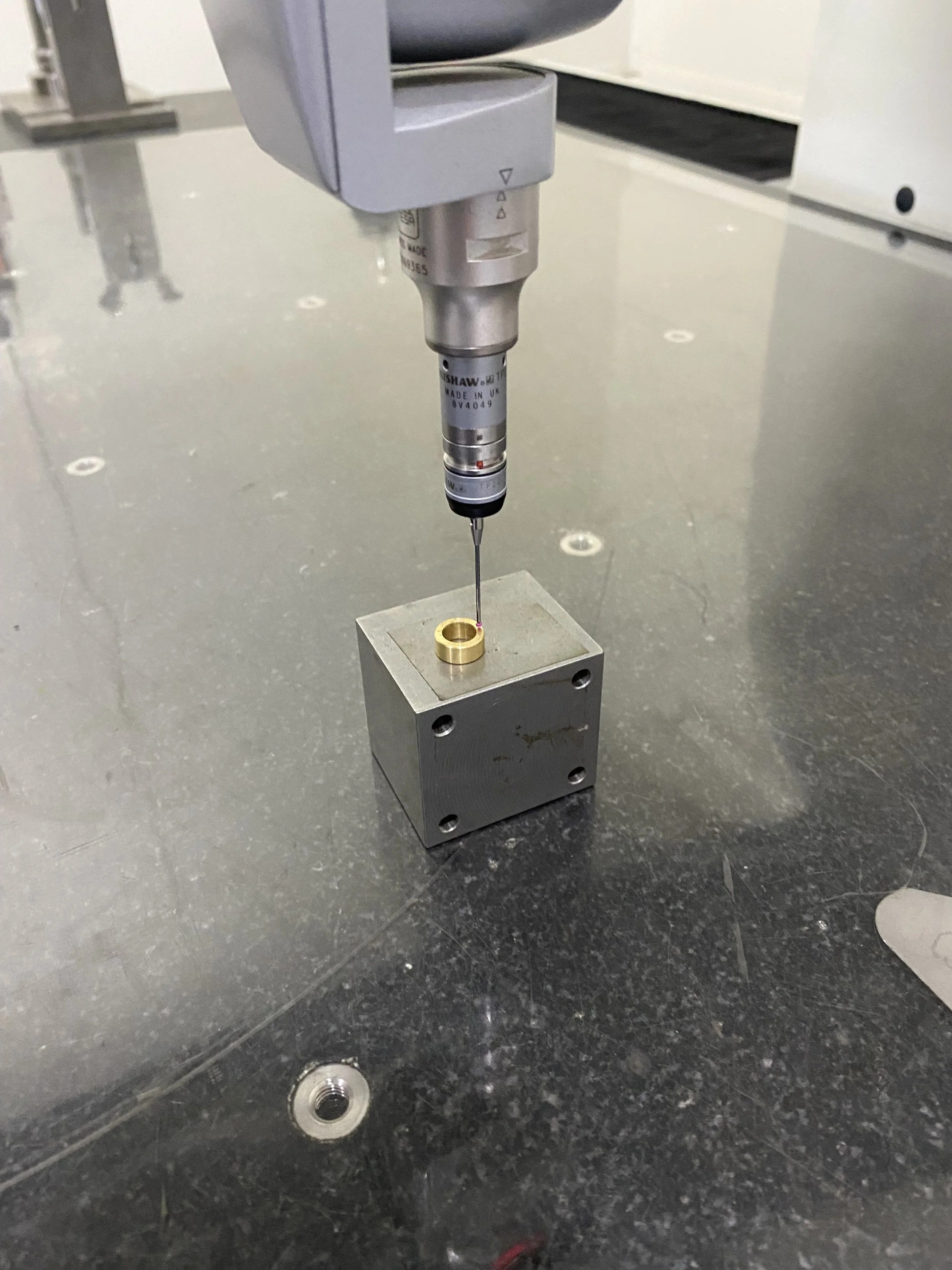 A coordinate measuring machine (CMM) is measuring a small gold-colored ring placed on a metallic square block on a black work surface.