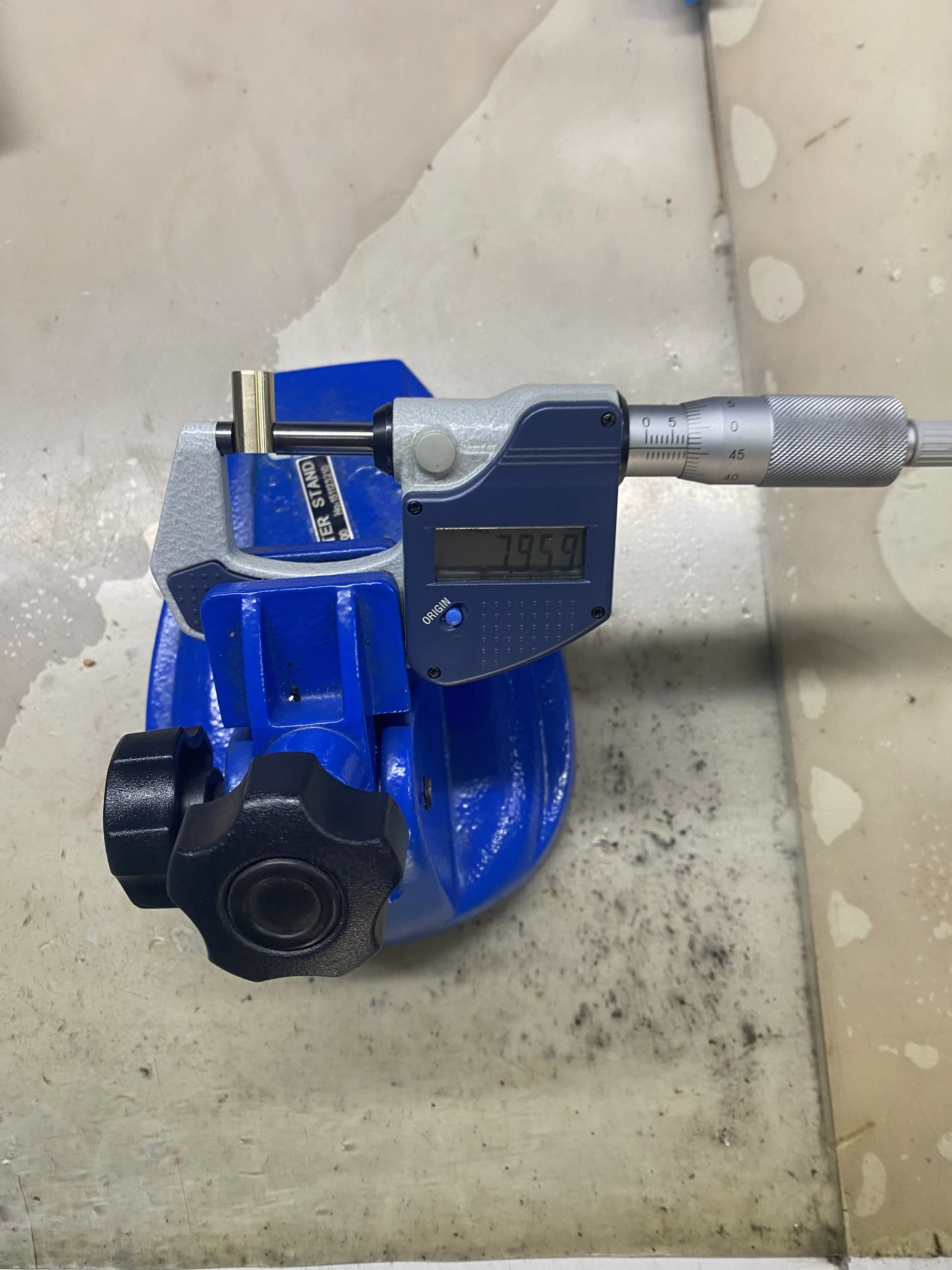 A digital caliper measuring the diameter of a round object with a reading of 7.959 millimeters, placed on a blue mechanical part on a work surface.