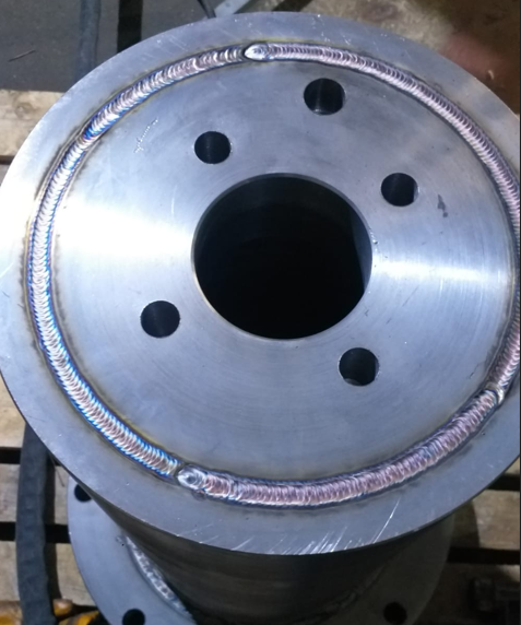 Close-up of a metal pipe flange with a welded bead around the edge, mounted on a wooden surface.